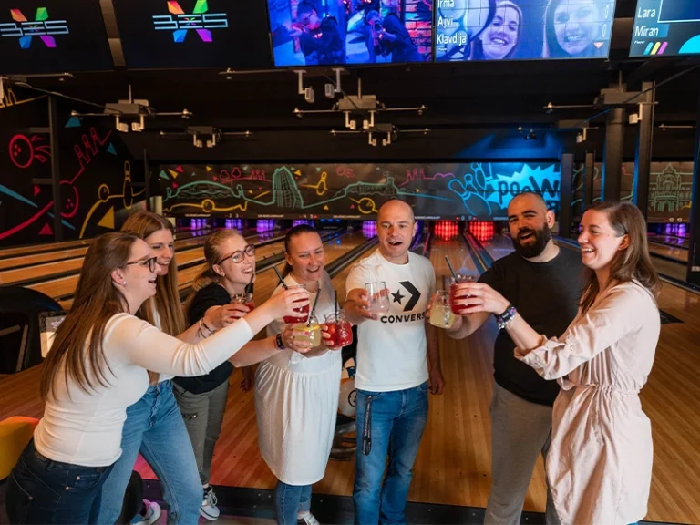 Gruppe von Menschen hebt Getränke in einer Bowling, umgeben von bunten Lichtern und Bildschirmen. Spaß und Teamgeist.