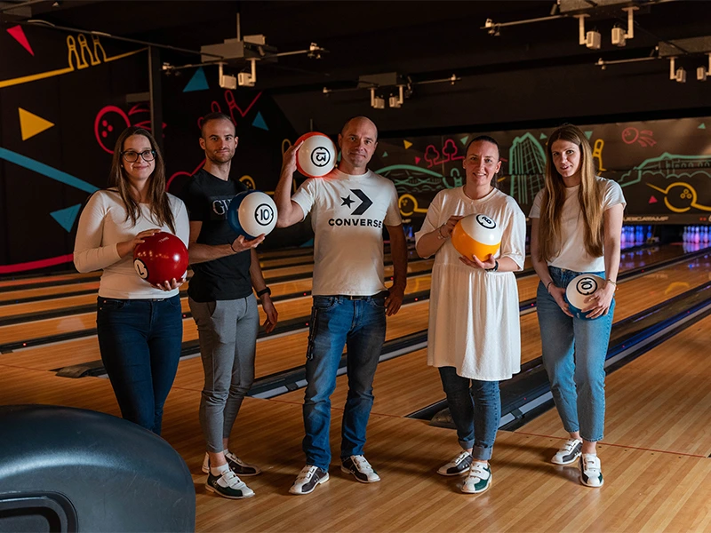 Gruppe von fünf Personen in einem Bowling, die Bowlingkugeln halten und Spaß haben. Bunte Beleuchtung und Bahnen im Hintergrund.