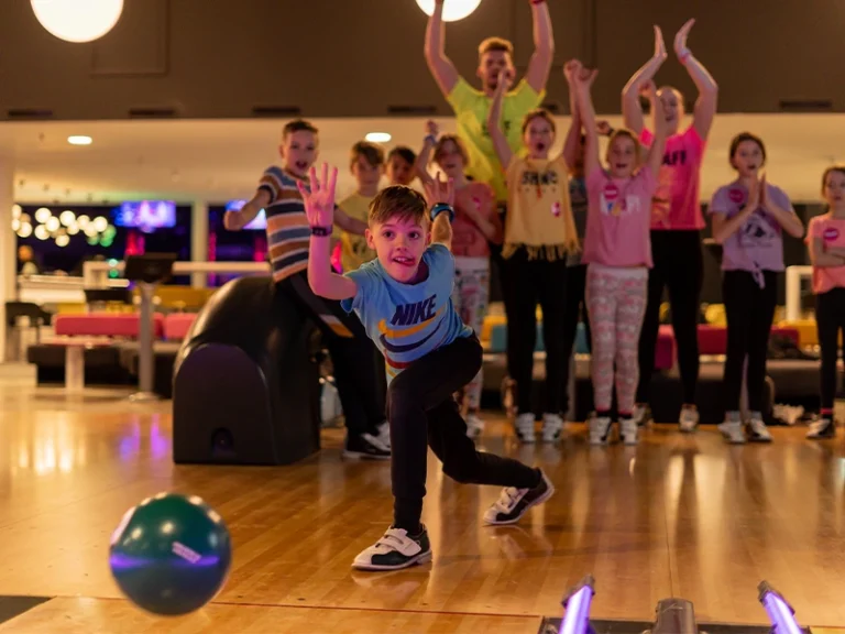 Ein Junge wirft eine Bowlingkugel in einer bunten Bowlingbahn, während Freunde jubeln und anfeuern.