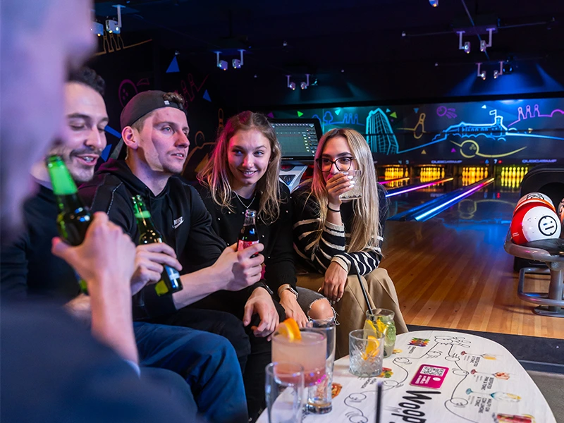 Freunde genießen Drinks und Unterhaltung in einer modernen Bowlingbahn mit bunten Lichtern und gemütlicher Atmosphäre.