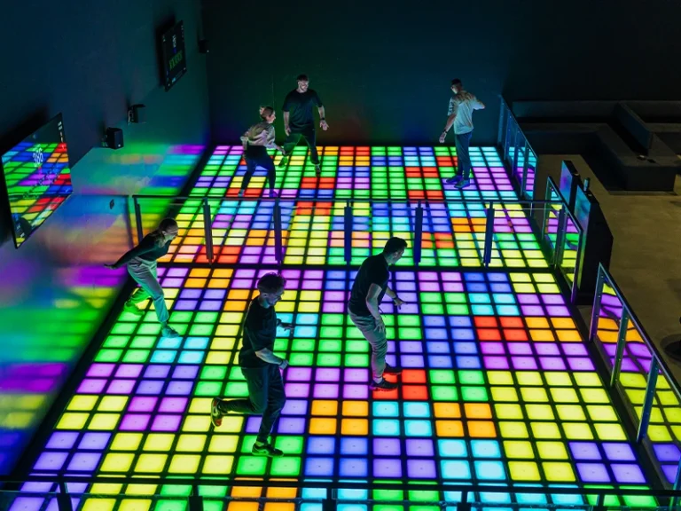 Tanzende Menschen auf einer bunten, leuchtenden Tanzfläche in einem modernen Club. Farbige Lichtspiele schaffen eine lebhafte Atmosphäre.