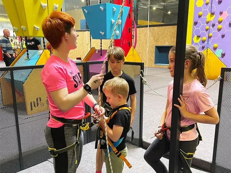 Eine Klettertrainerin zeigt Kindern, wie sie sich für das Klettern sichern. Indoor-Kletterhalle mit bunten Wänden.