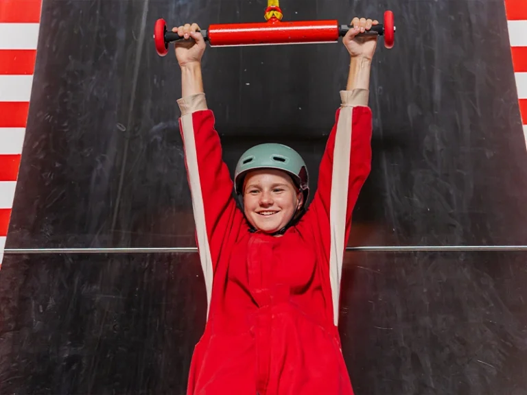 Kind in rotem Anzug und Helm hängt an einer Stange in einer Kletterhalle. Spaß und Abenteuer beim Klettern.