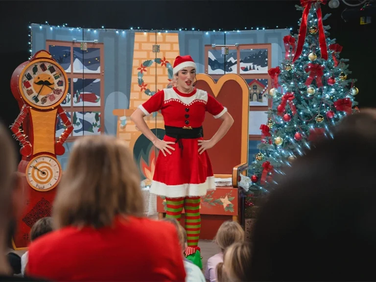 Ein Weihnachtsmann in einem festlichen Kostüm unterhält Kinder während einer Weihnachtsaufführung. Bunte Dekorationen im Hintergrund.
