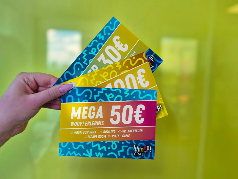Händehaltung von drei Gutscheinen im Wert von 30€, 100€ und einem Mega-Gutschein über 50€ vor einer hellgrünen Wand.