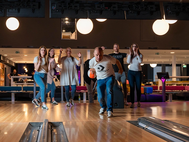 Gruppe von Menschen beim Bowling in einer modernen Bowlingbahn, während ein Mann mit einer Bowlingkugel im Begriff ist, seinen Wurf zu machen.