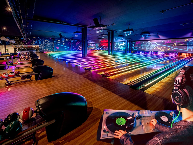 Blick auf einen modernen Club mit bunten Lichtern und einem DJ, der auf einem Plattenspieler auflegt. Die Bowlingbahn ist beleuchtet und lädt zum Feiern und Spaß haben ein.