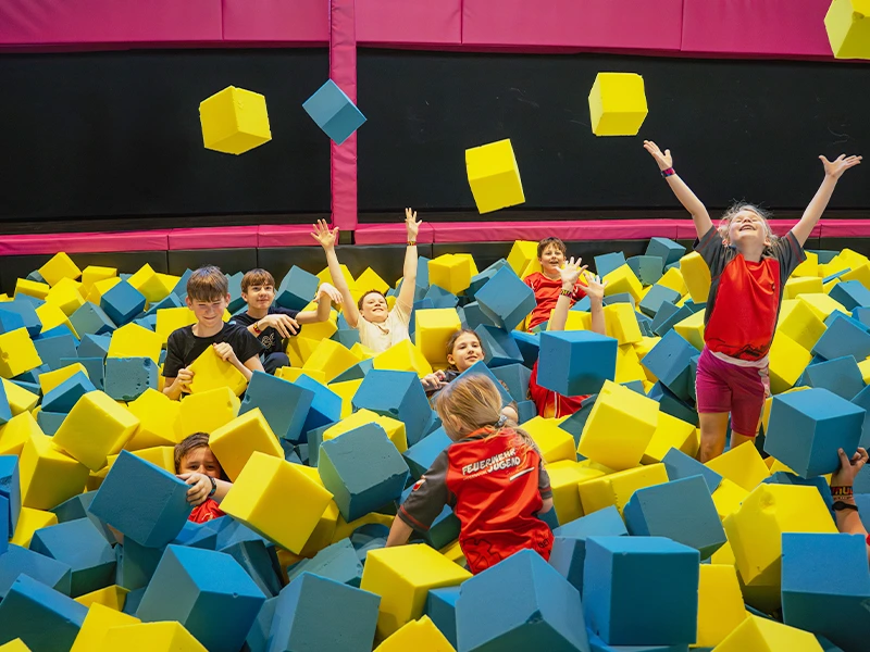 Kinder spielen in einem Trampolinpark und werfen bunte Schaumstoffwürfel in die Luft. Spaß und Aktivität im Vordergrund.