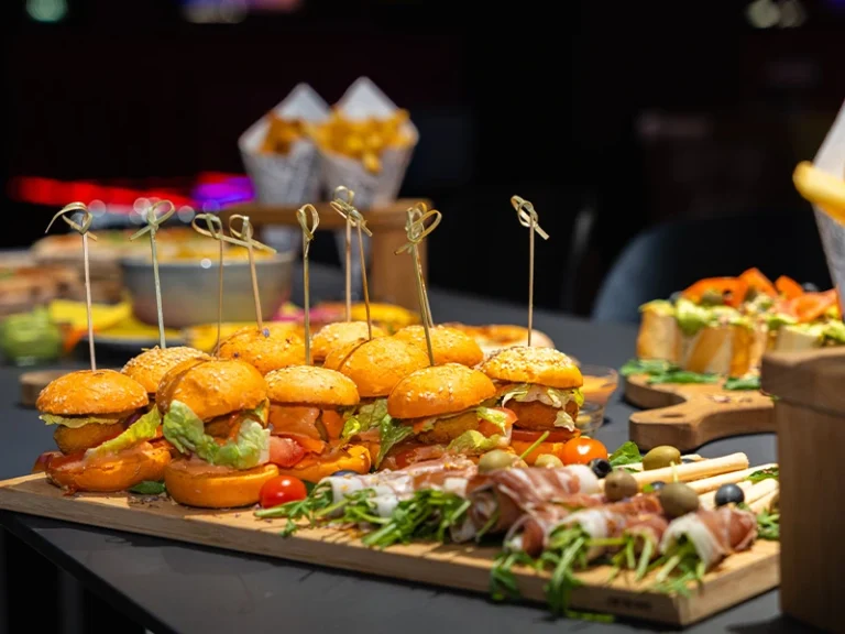 Leckere Mini-Burger auf einem Holzbrett, umgeben von Snacks und frischen Zutaten, ideal für Partys und Feiern.