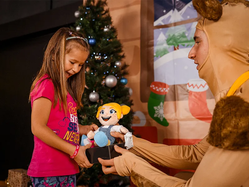 Ein Mädchen erhält ein Plüschtier von einem verkleideten Charakter vor einem Weihnachtsbaum. Freude und Überraschung stehen im Vordergrund.