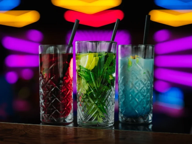 Drei bunte Cocktails in Gläsern mit Strohhalmen vor einem leuchtenden, farbenfrohen Hintergrund. Ideal für Partys.