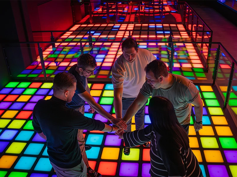 Gruppe von fünf Personen auf einem bunten, beleuchteten Tanzboden, die sich zum Teamwork versammeln.