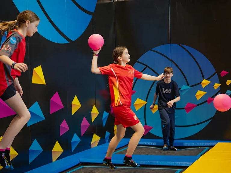 Kinder spielen in einer Trampolinhalle. Ein Mädchen wirft einen pinken Ball, während andere aktiv sind.